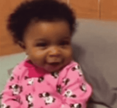 A Baby Girl In A Pink Pajama Set Is Smiling While Sitting On A Bed . GIF