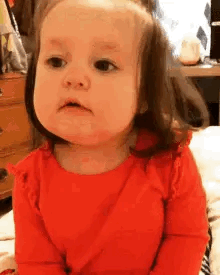 A Baby Girl In A Red Shirt Is Sitting On A Bed Making A Funny Face . GIF
