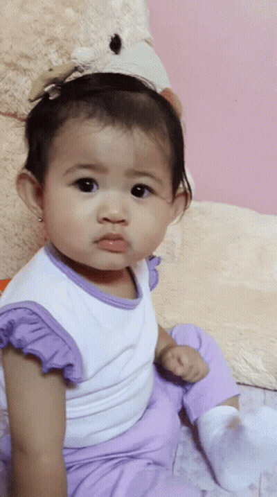 A Baby Girl Wearing Purple Pants And A White Shirt Is Sitting Next To A Teddy Bear GIF