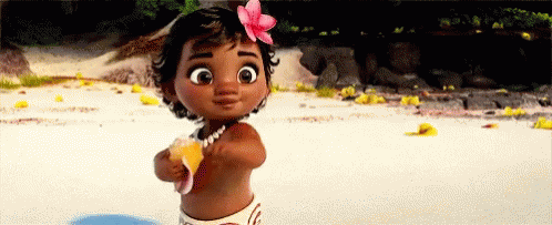 A Baby Girl With A Flower In Her Hair Is Standing On The Beach Holding A Shell . GIF