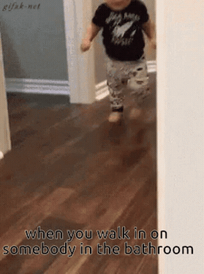 A Baby In A Black Shirt Is Walking On A Wooden Floor In A Bathroom . GIF