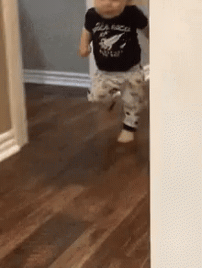 A Baby In A Black Shirt Is Walking On A Wooden Floor GIF