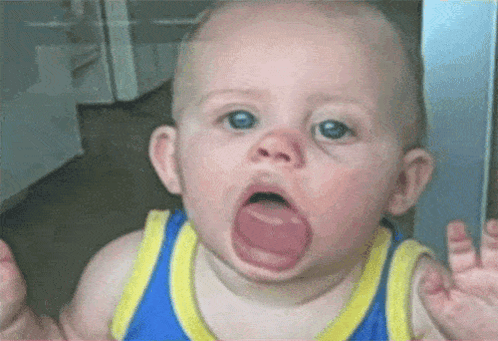 A Baby In A Blue And Yellow Tank Top Is Making A Funny Face With His Mouth Open . GIF