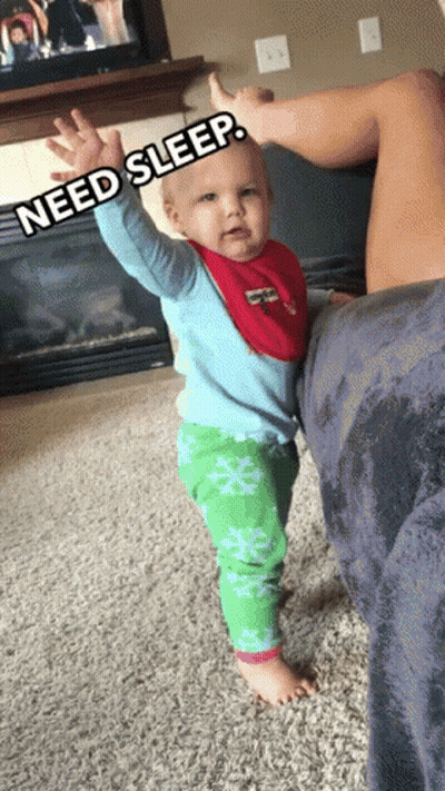 A Baby In A Blue Shirt And Green Pants Is Standing Next To A Woman Who Is Laying On A Couch GIF