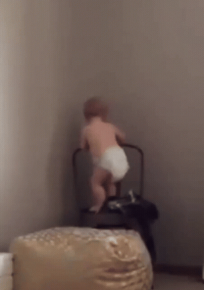 A Baby In A Diaper Is Standing Next To A Chair . GIF