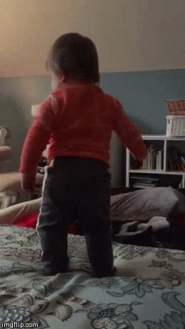 A Baby In A Red Shirt Is Standing On A Bed In A Living Room . GIF
