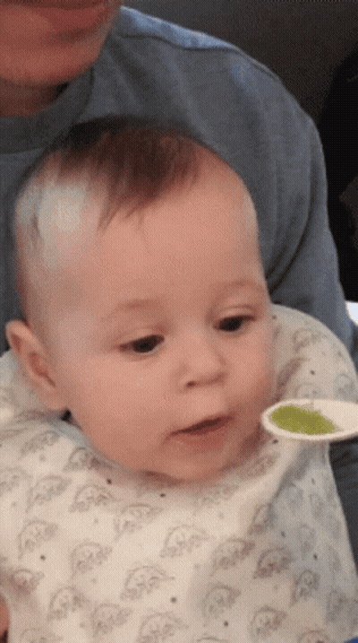 A Baby Is Being Fed With A Spoon By A Man In A Blue Shirt GIF