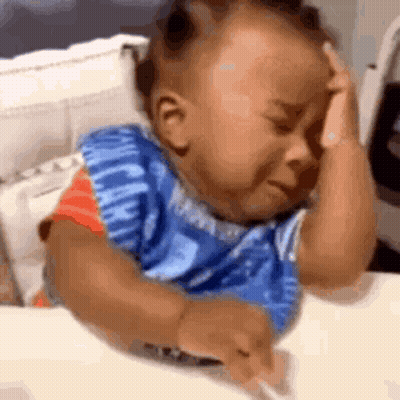 A Baby Is Crying While Sitting At A Table With His Hands On His Head . GIF