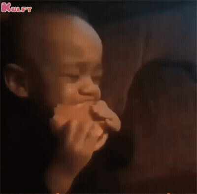A Baby Is Eating A Hamburger In A Dark Room While A Woman Looks On . GIF