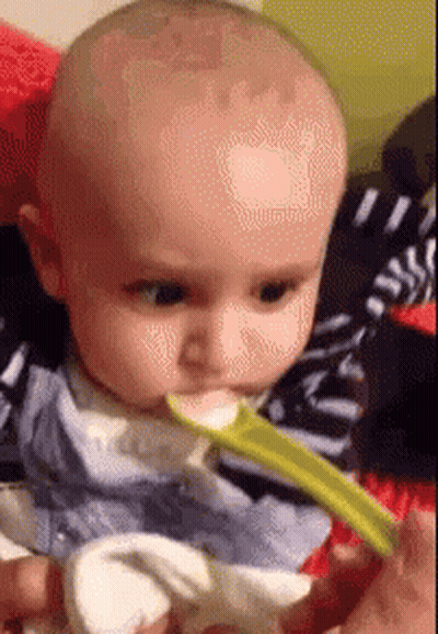 A Baby Is Eating Food With A Spoon And Looking At The Camera . GIF