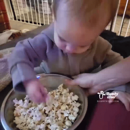 A Baby Is Eating Popcorn From A Bowl With A Fork GIF