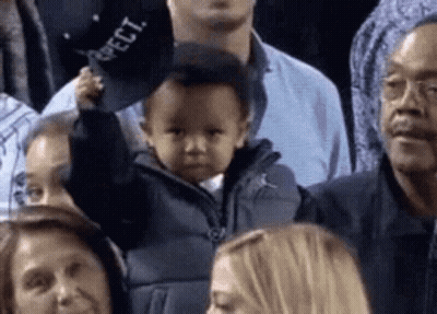 A Baby Is Holding A Hat In A Crowd Of People At A Basketball Game . GIF