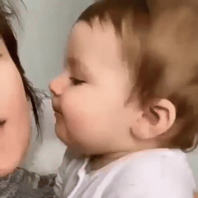 A Baby Is Kissing A Woman On The Cheek And Looking At The Camera . GIF