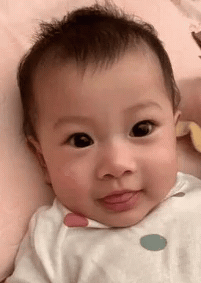 A Baby Is Laying On A Bed And Looking At The Camera With His Tongue Out . GIF