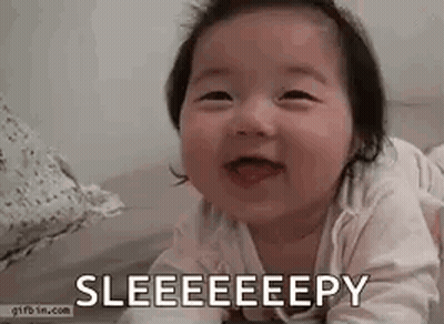 A Baby Is Laying On Its Stomach On A Bed And Smiling With Its Tongue Out . GIF