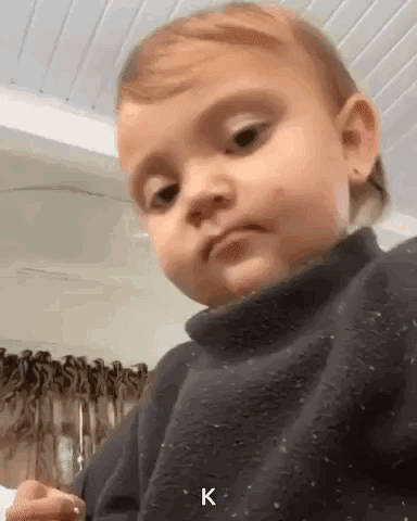 A Baby Is Making A Funny Face And Looking At The Camera While Wearing A Black Sweater . GIF