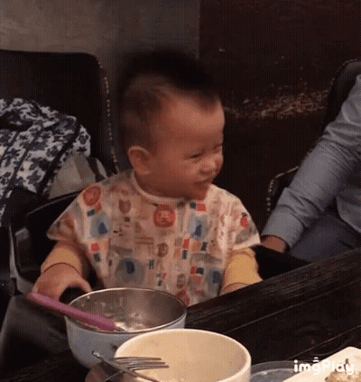 A Baby Is Sitting At A Table With A Bowl Of Food And A Spoon And Fork GIF
