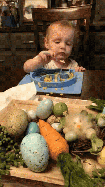 A Baby Is Sitting At A Table With A Plate Of Food And A Spoon GIF