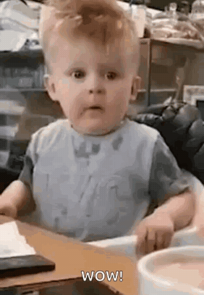 A Baby Is Sitting In A High Chair And Making A Funny Face While Looking At The Camera . GIF