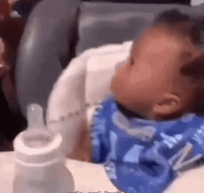 A Baby Is Sitting In A High Chair With A Bottle GIF