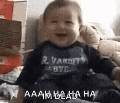 A Baby Is Sitting On A Bed And Laughing While Wearing A Varsity Shirt . GIF