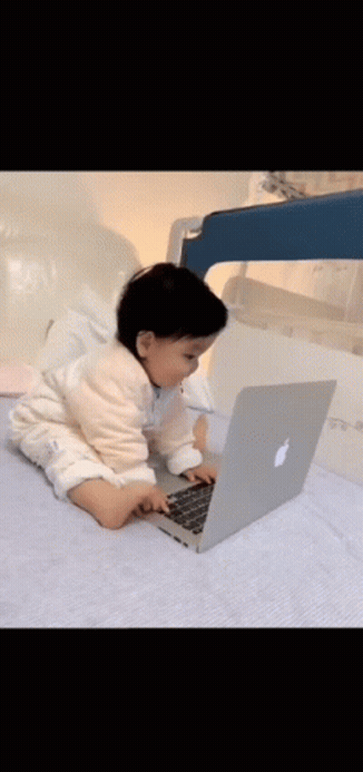 A Baby Is Sitting On A Bed Using An Apple Laptop Computer . GIF