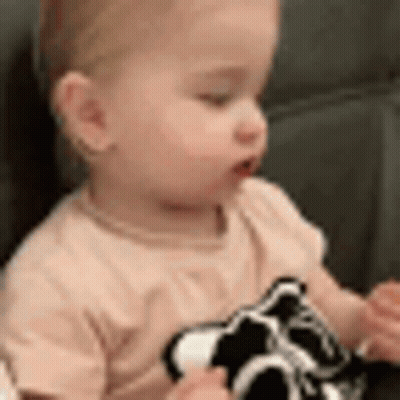 A Baby Is Sitting On A Couch Holding A Black And White Toy . GIF