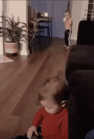 A Baby Is Sitting On A Couch In A Living Room While A Little Boy Stands In The Background . GIF