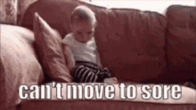A Baby Is Sitting On A Couch With The Words `` Can 'T Move To Sore '' Written On The Bottom . GIF