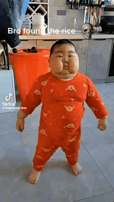 A Baby Is Standing In A Kitchen Wearing A Red Pajama Set . GIF
