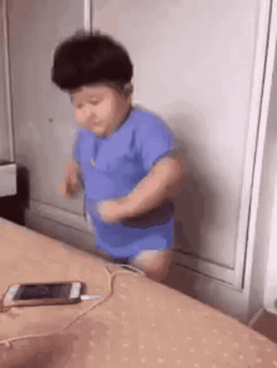 A Baby Is Standing On A Bed Next To A Cell Phone . GIF
