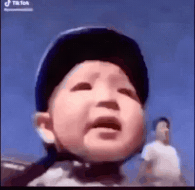 A Baby Is Wearing A Helmet And Crying While A Man Stands Behind Him . GIF