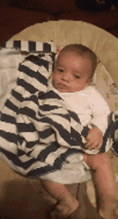 A Baby Is Wrapped In A Striped Blanket And Sitting In A Bouncer . GIF