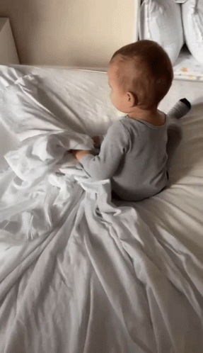 A Baby Laying On A Bed With A White Blanket GIF