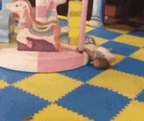 A Baby Laying On A Checkered Floor Next To A Pink Rocking Horse . GIF