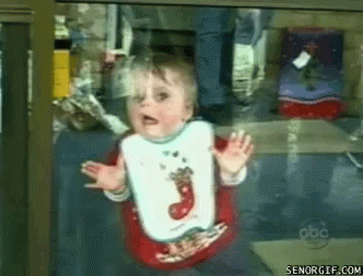 A Baby Wearing A Bib With A Christmas Stocking On It Behind A Glass Door GIF