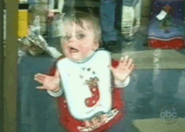 A Baby Wearing A Bib With A Christmas Stocking On It Is Standing In Front Of A Abc Sign GIF