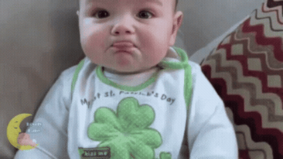 A Baby Wearing A Bib With A Clover On It Is Making A Funny Face While Sitting On A Couch . GIF