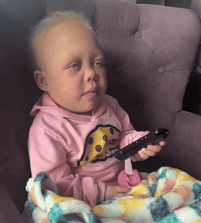 A Baby Wearing A Pink Shirt With Cheese On It Holds A Remote Control GIF