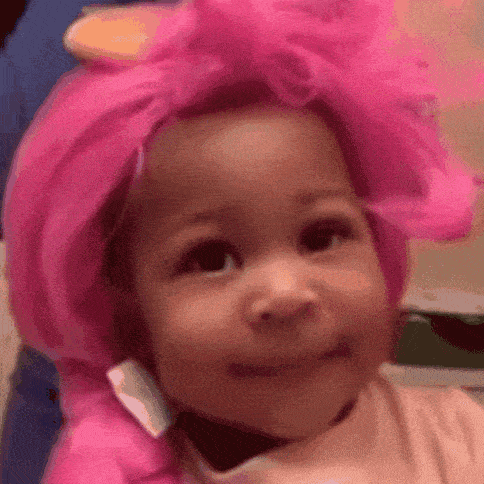 A Baby Wearing A Pink Wig And A Pink Hat Is Smiling . GIF