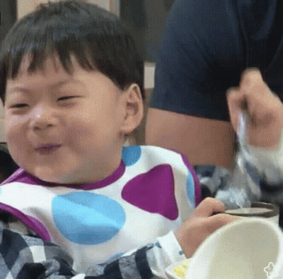 A Baby Wearing A Polka Dot Bib Is Smiling While Eating From A Bowl . GIF