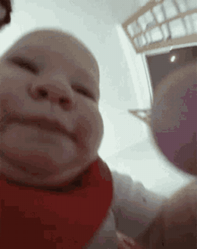 A Baby Wearing A Red Bib Is Looking At The Camera And Smiling . GIF