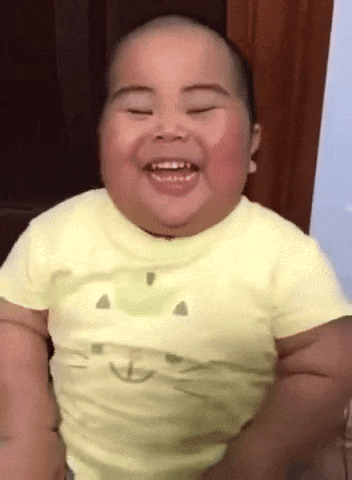 A Baby Wearing A Yellow Shirt With A Smiley Face On It Is Smiling GIF