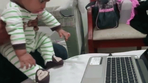 A Baby Wearing Monkey Slippers Is Sitting On A Laptop GIF