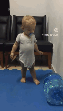 A Baby With A Pacifier In His Mouth Is Walking On A Blue Mat GIF