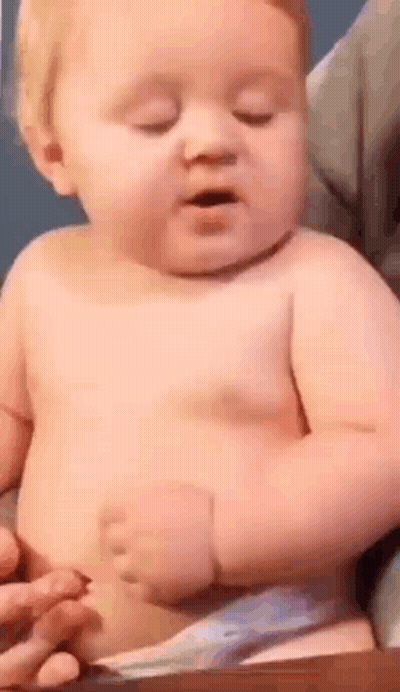 A Baby Without A Shirt Is Sitting On A Table With His Eyes Closed And His Mouth Open . GIF