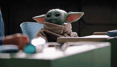 A Baby Yoda Is Sitting At A Table With A Scarf Around His Neck . GIF