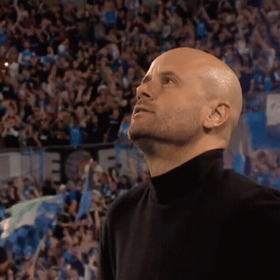 A Bald Man In A Black Turtleneck Stands In Front Of A Crowd And Looks Up GIF