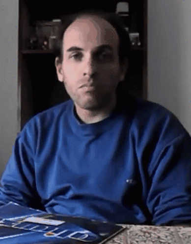 A Bald Man In A Blue Shirt Is Sitting At A Table With A Magazine On It GIF