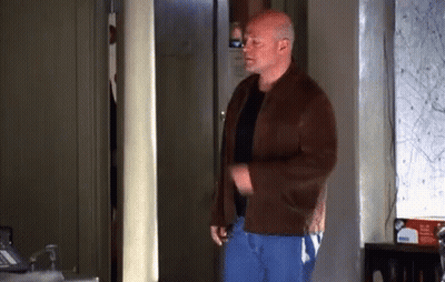 A Bald Man In A Brown Jacket And Blue Pants Is Standing In A Room . GIF